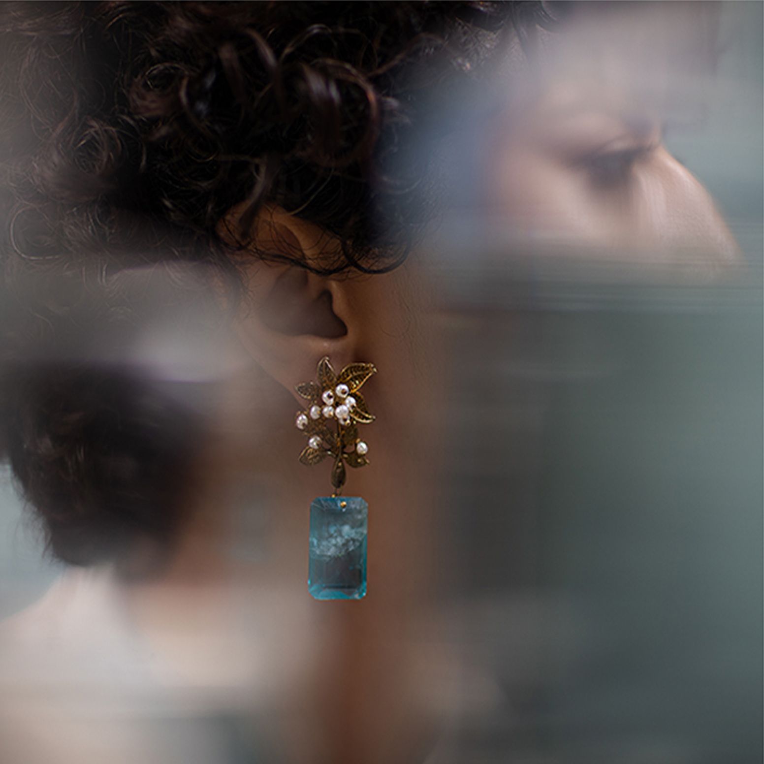 Close-up of an ornate earring with floral gold details, pearls, and a large blue gemstone, photographed through soft-focus glass for an artistic advertising concept.