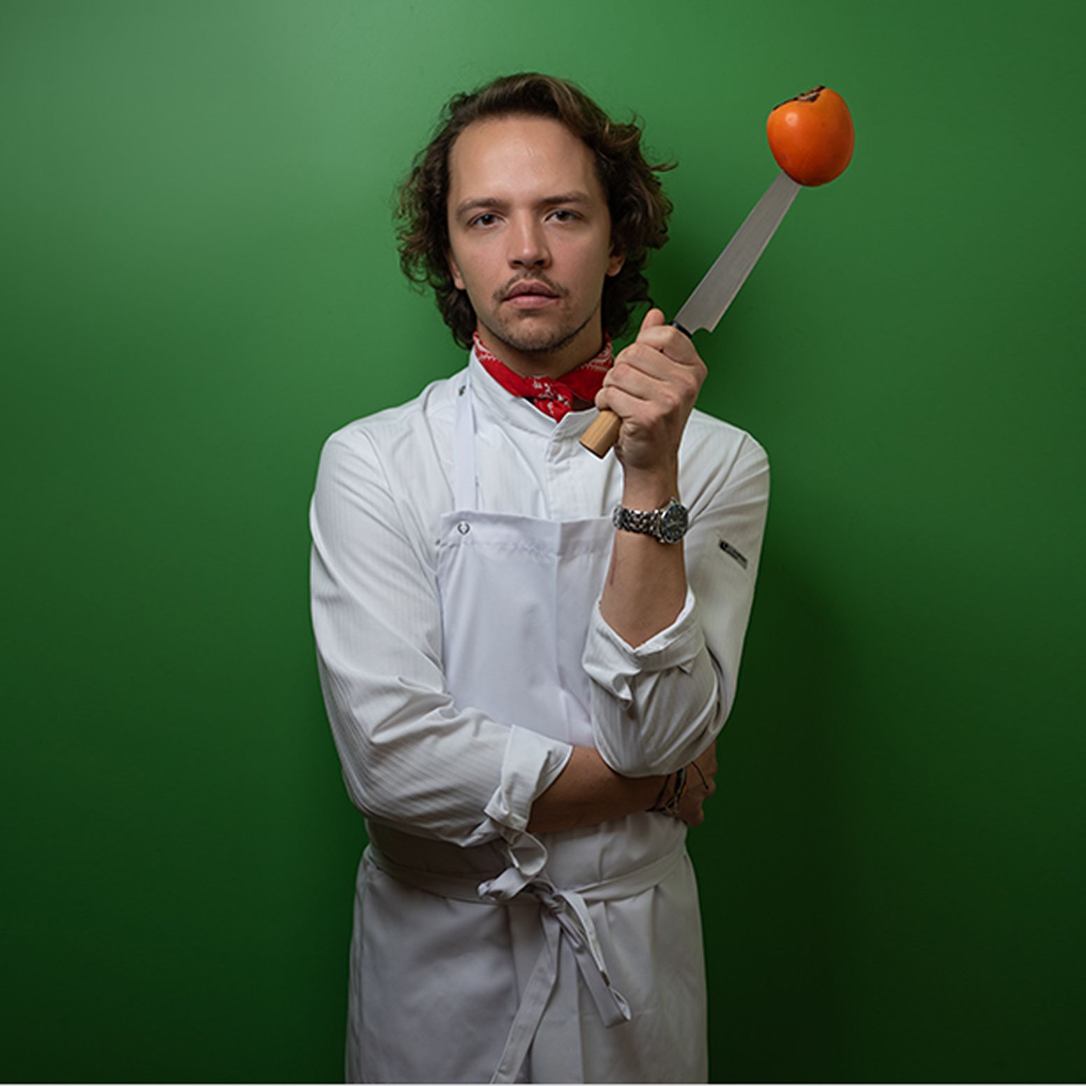 Chef in a white uniform poses confidently with arms crossed, holding a knife balancing a persimmon, set against a vibrant green backdrop—blending corporate portraiture with personality.