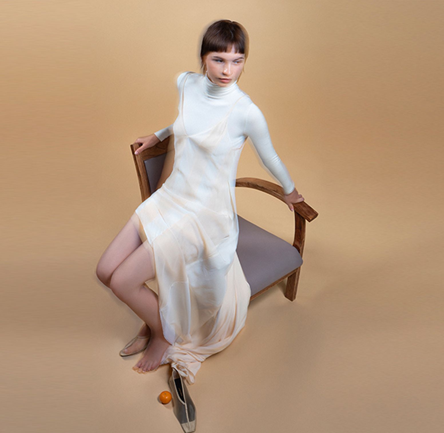 A young woman in a sheer, modern dress poses elegantly on a wooden chair against a monochrome beige background, blending contemporary style with minimalist fashion aesthetics.
