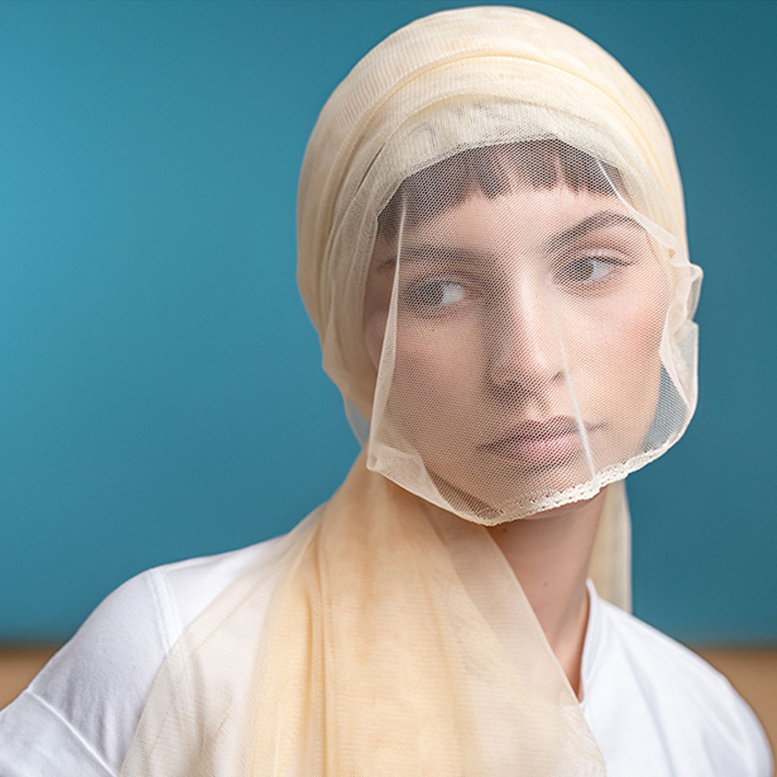 Soft-lit portrait of a young woman gazing away while wearing a translucent beige veil over her head, set against a cyan background—conveying mystery and elegance.